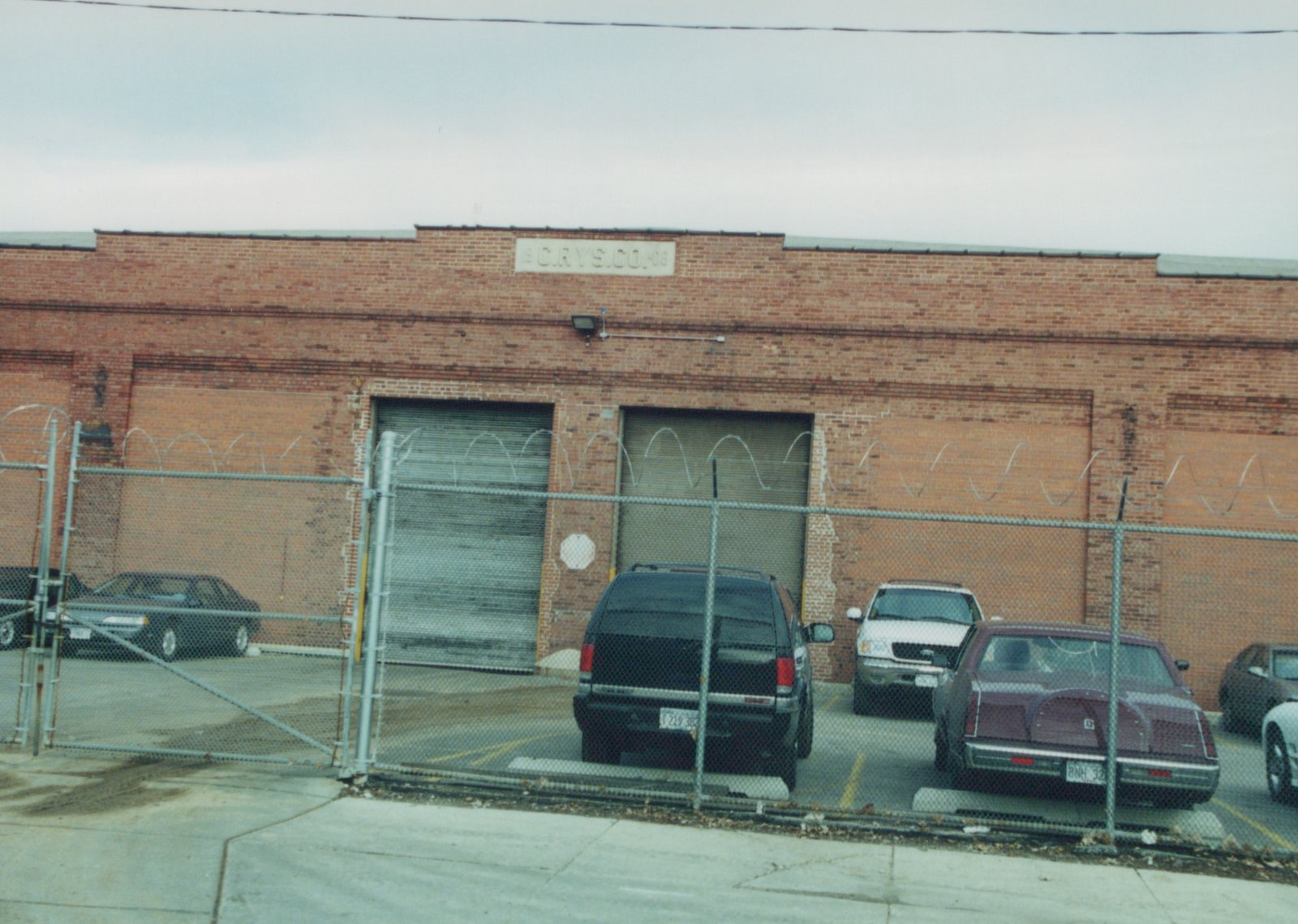 A brick building with a sign that reads 'C.R.Y.S.CO.' is seen behind a chain-link fence with barbed wire; several cars are parked in the lot in front of it on an overcast day.
