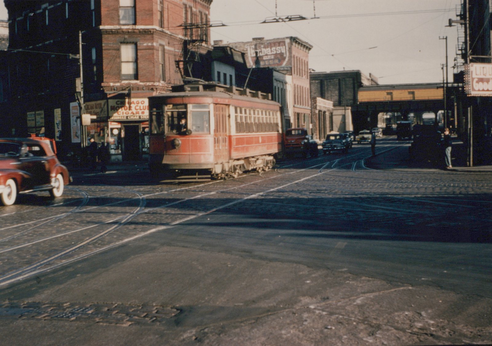 A red and cream streetcar from the Chicago Surface Lines turns at a street intersection in Chicago, surrounded by 1940s automobiles, brick buildings, and visible trolley wires.