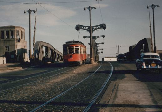 A red and white Chicago Surface Lines streetcar numbered 141 is positioned on a bascule bridge with vintage cars and a control tower nearby under a clear sky.