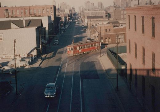 A high-angle view of a red streetcar turning a corner on a city street, surrounded by 1940s cars and brick buildings, with a person standing on the car's roof.