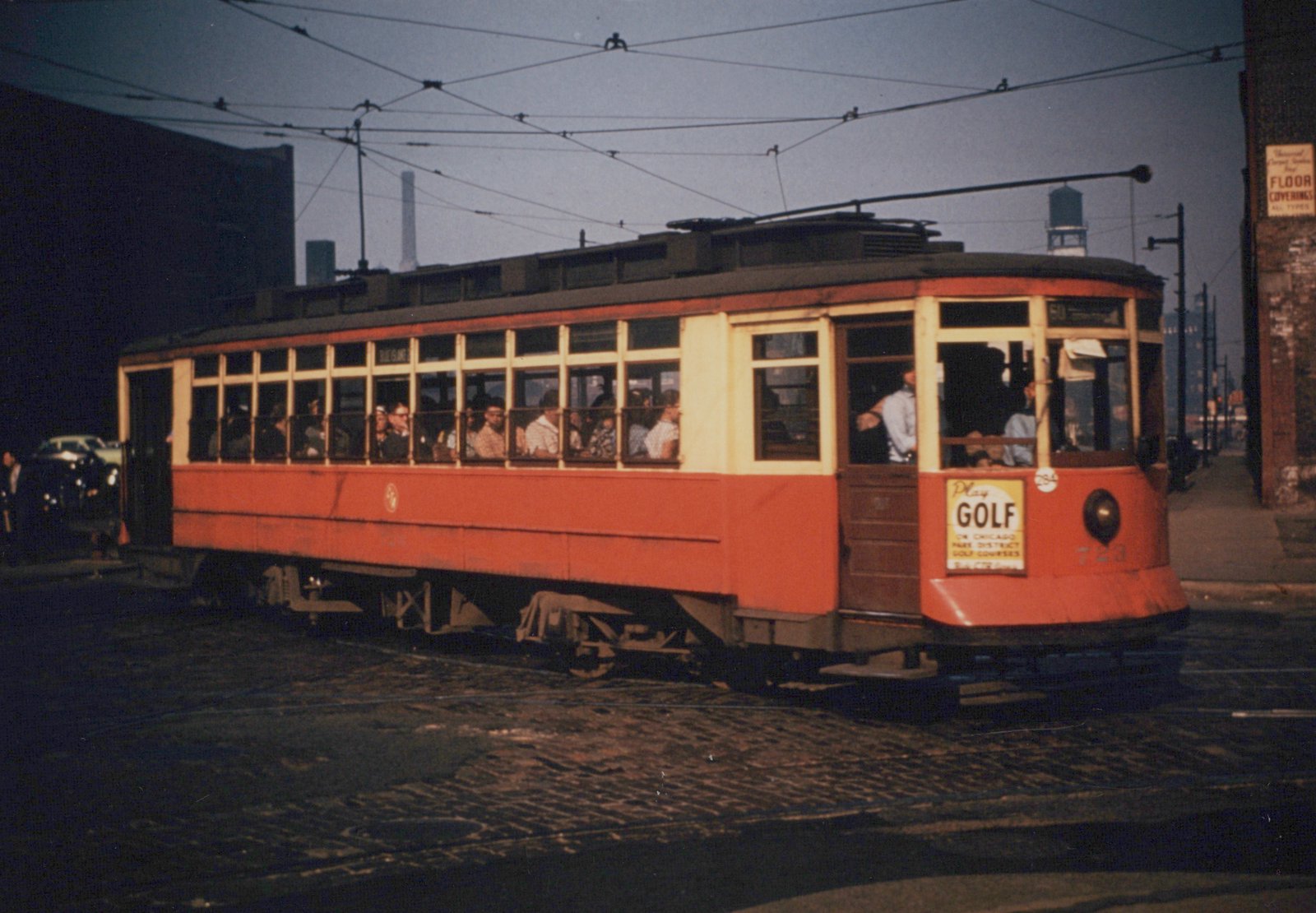 A vintage red and cream streetcar numbered 723, with a destination sign reading 'THE ISLAND', is shown on a cobblestone street with passengers visible through its windows, under overhead wires and next to a building with a 'FLOOR COVERINGS' sign.