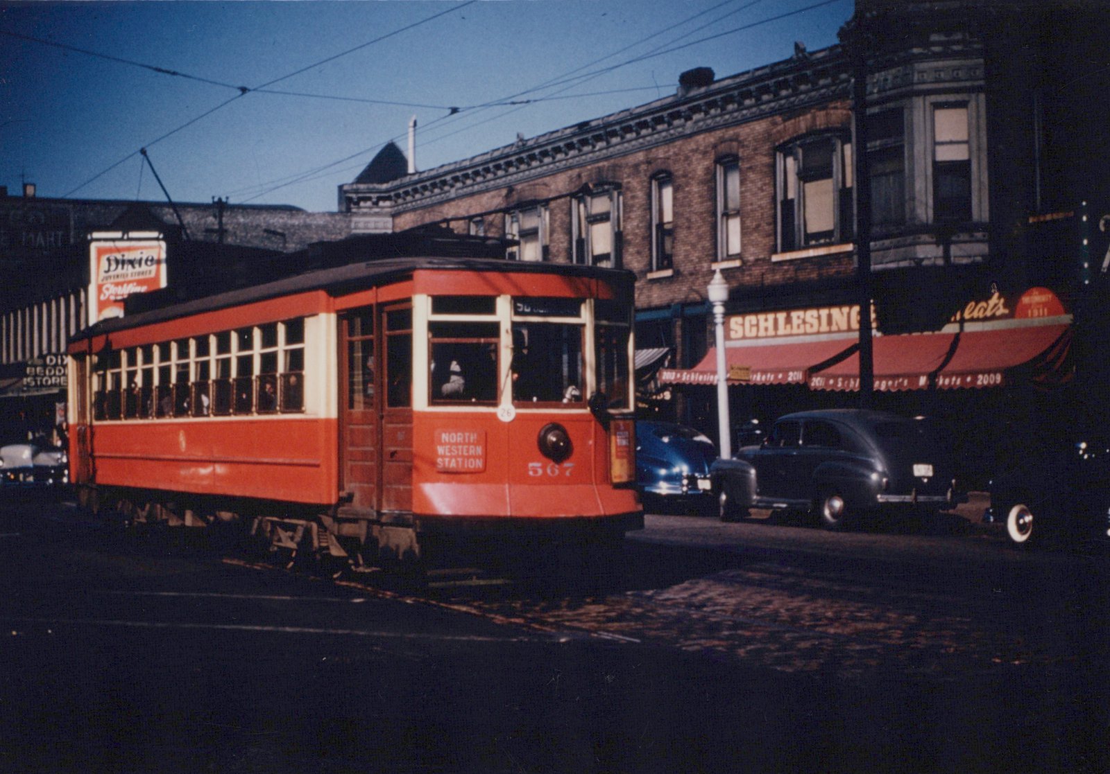 A red and cream streetcar with the number 567 and 'NORTH WESTERN STATION' shown on its front is stopped on a city street with cars and brick buildings in the background.