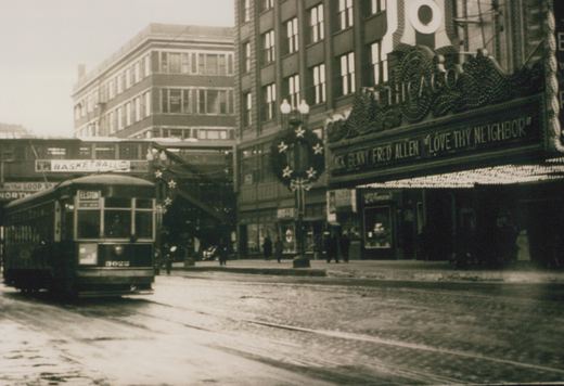 A vintage black-and-white photo showing a streetcar on a wet city street in front of a movie theater, with pedestrians and an elevated train structure in the background.