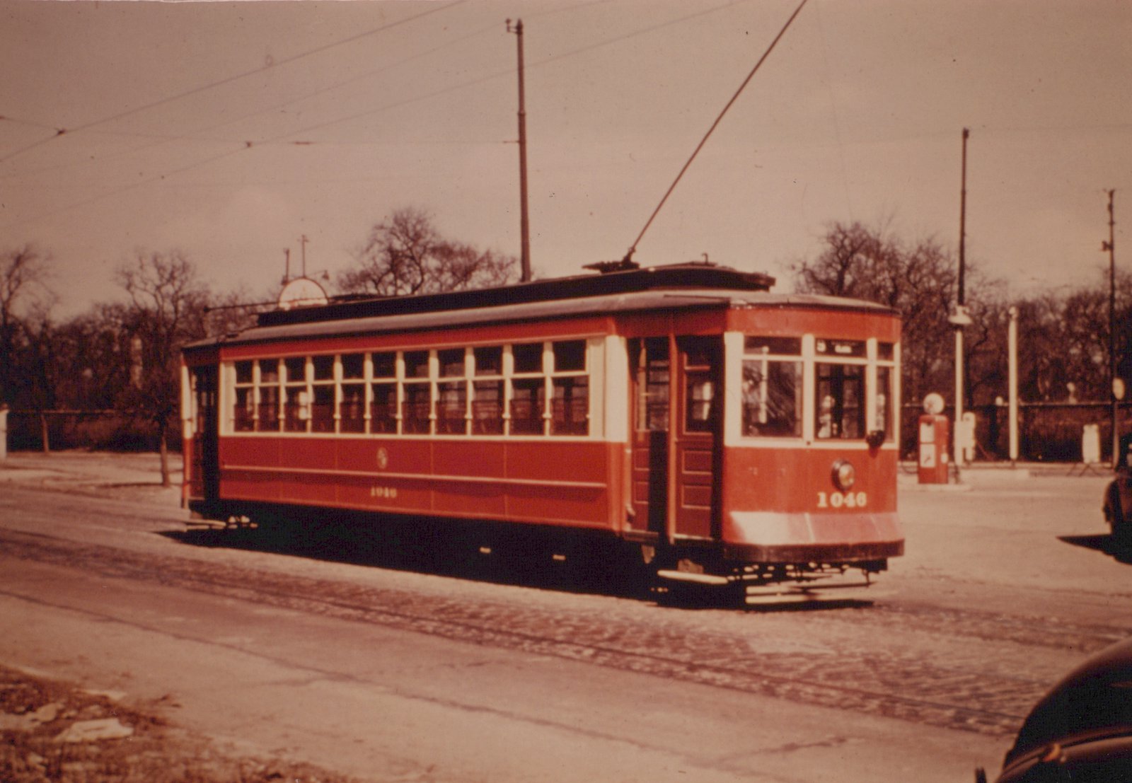 A red and cream streetcar numbered 1046 is shown from a three-quarter front view, running on a street with bare trees and a gasoline pump in the background.