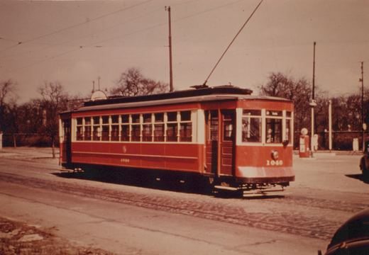 A red and cream streetcar numbered 1046 is shown from a three-quarter front view, running on a street with bare trees and a gasoline pump in the background.