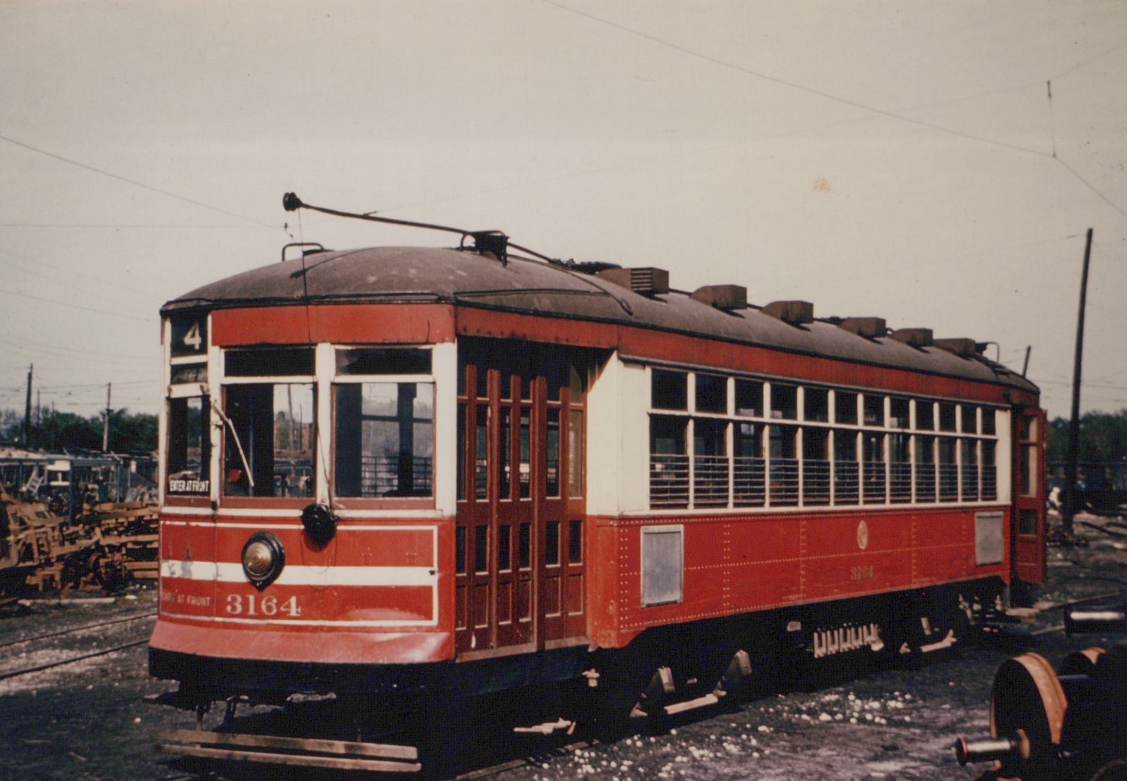 A red and cream streetcar numbered 3164 is parked in a rail yard, with overhead wires visible and other scrapped cars in the background.