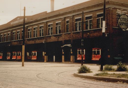 A row of red Birney streetcars is parked inside a large brick carbarn with the number 5, 6, and 7 painted above the bays; a 'CITIES SERVICE' sign is visible on the building's corner.