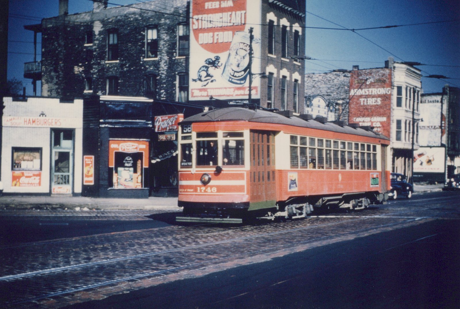 A vintage orange and cream streetcar with the number 1746 and route 58 sign, moving down a city street lined with 1930s-era storefronts and billboards under a clear blue sky.
