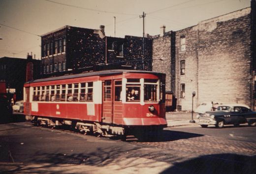A vintage red streetcar with the number 556 turns a corner on a city street, with brick buildings and a classic automobile in the background.
