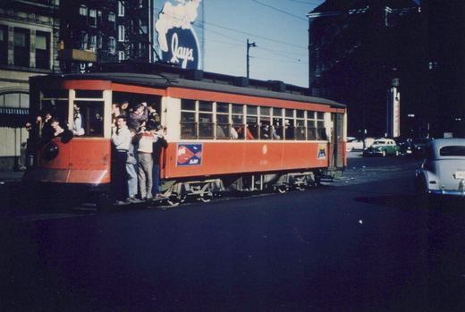 A red and cream Chicago Surface Lines streetcar, number 436, is stopped on a city street with several passengers standing on its running board, surrounded by vintage 1940s automobiles and a Lay's potato chips billboard.