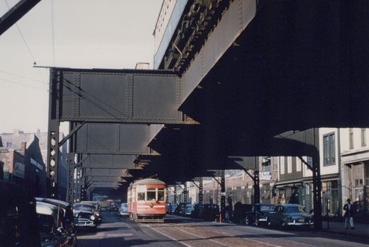 A red and cream streetcar marked CSL 1721 moves down a street beneath a dark steel elevated structure, flanked by parked 1940s cars and a few pedestrians.