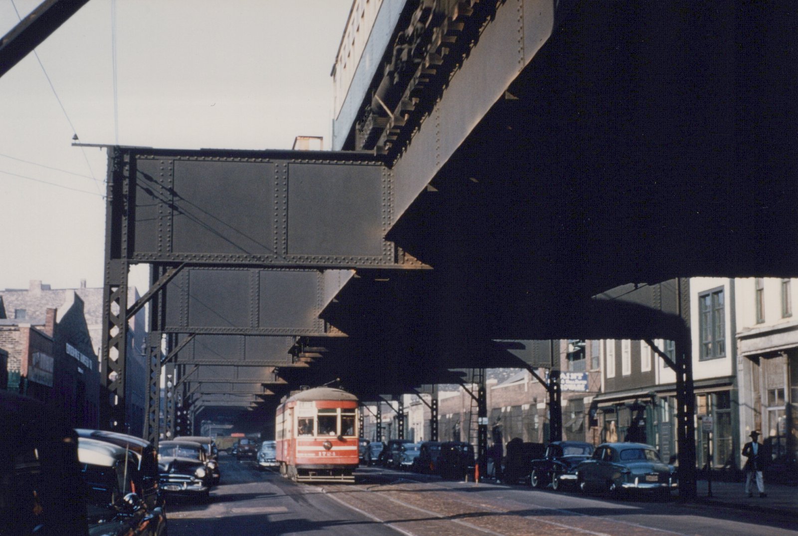 A red and cream streetcar marked CSL 1721 moves down a street beneath a dark steel elevated structure, flanked by parked 1940s cars and a few pedestrians.