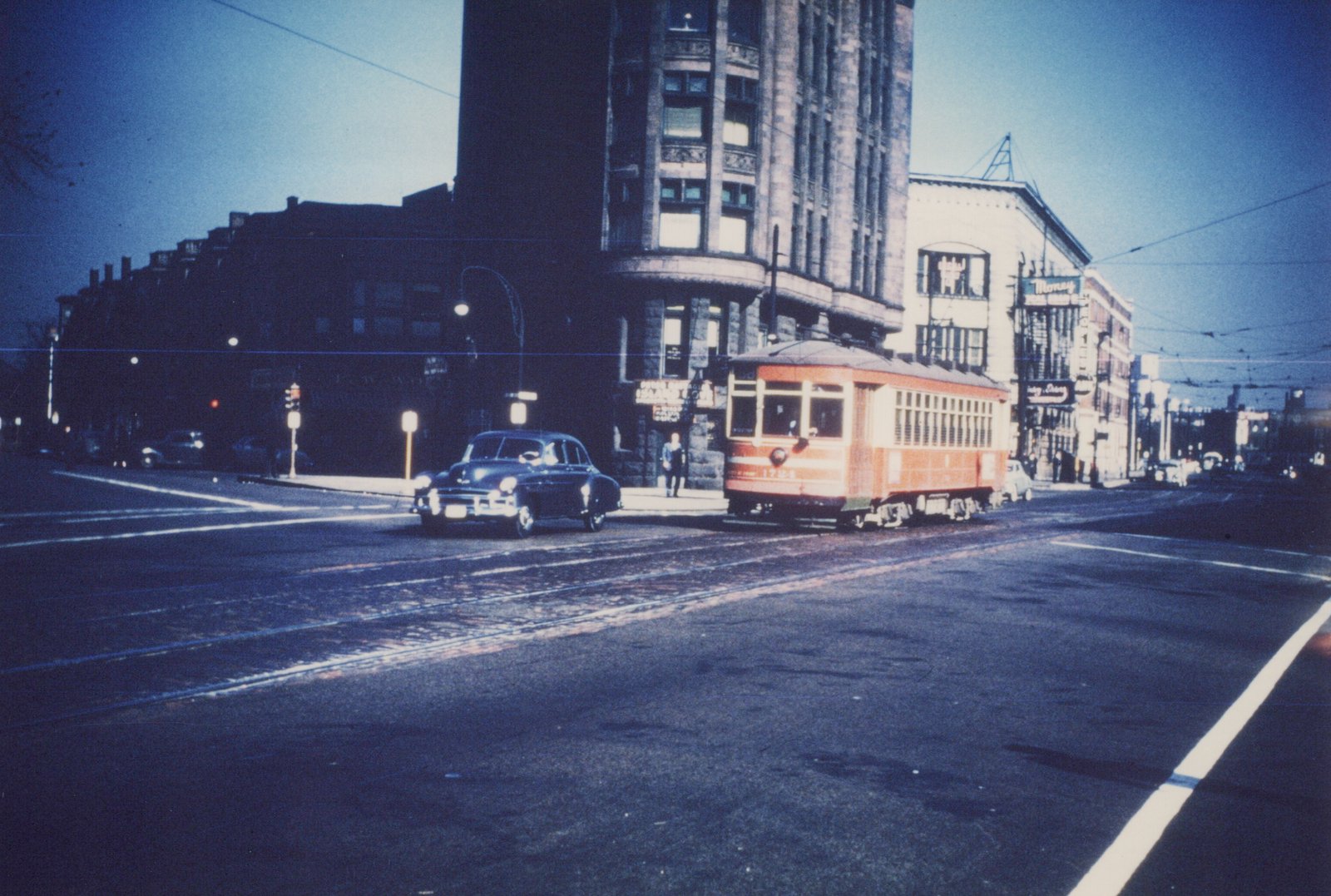A vintage orange streetcar on city streets at dusk, with a classic car and pedestrians nearby.