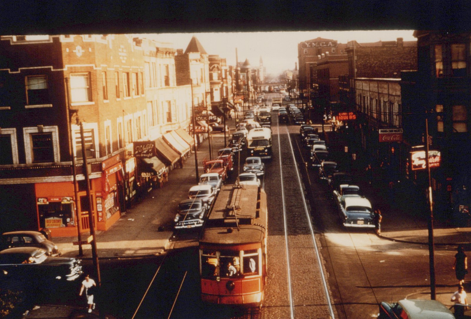 A red streetcar runs down the center of a bustling city street, surrounded by vintage cars and pedestrians under an elevated structure.