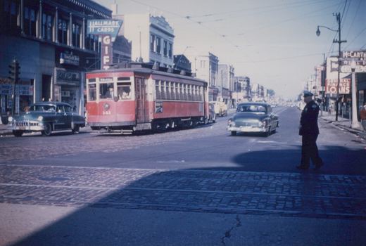 A red and cream streetcar with the number 141 travels down a city street lined with shops and cars from the late 1940s, while a police officer directs traffic.