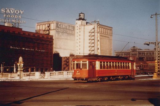 A red Birney streetcar, number 277, travels on a snowy city street under trolley wires, with large brick and white factory buildings behind it.