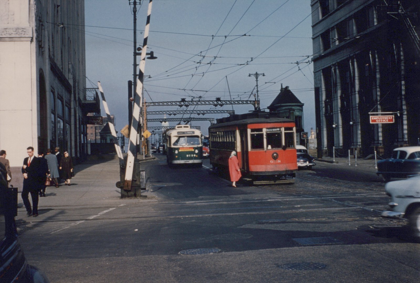 Two vintage Chicago streetcars, a green PCC and a red Peter Witt, are stopped at a grade crossing with automobiles and pedestrians nearby on a sunny day.