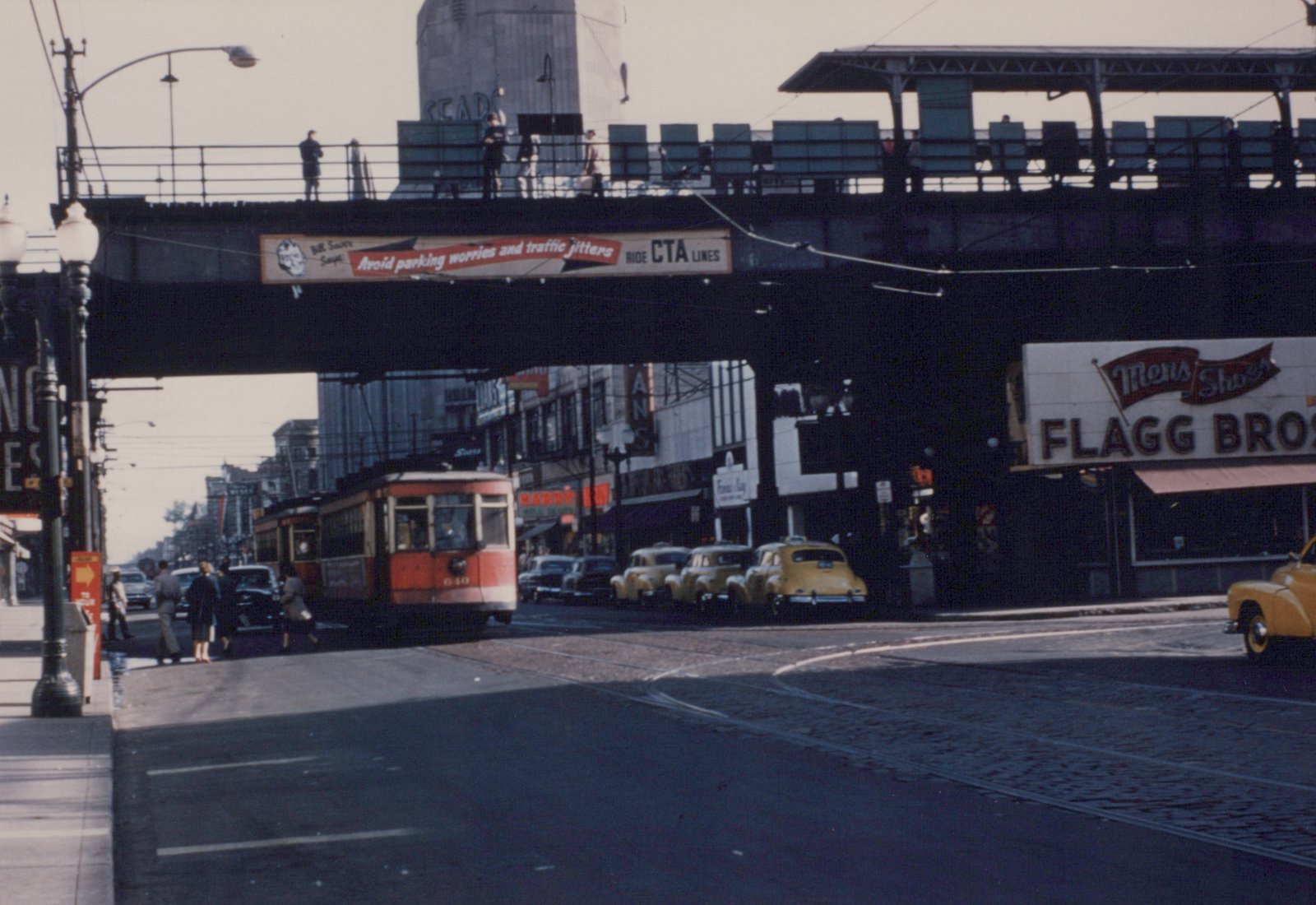 A red and cream streetcar with the number 640 travels down a street under a dark elevated structure, with people on the sidewalk and vintage cars parked along the curb.