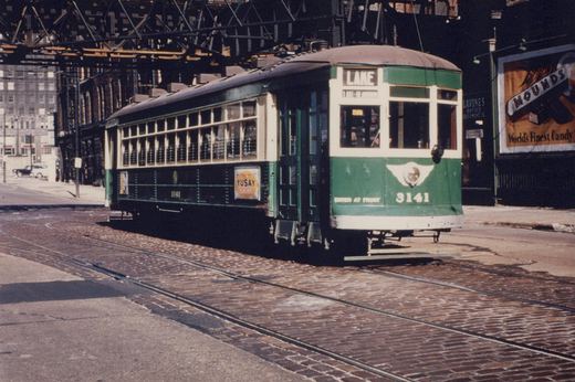 A vintage green and cream streetcar numbered 3141 with a 'LAKE' destination sign, parked on cobblestone tracks under a steel elevated structure in a city.