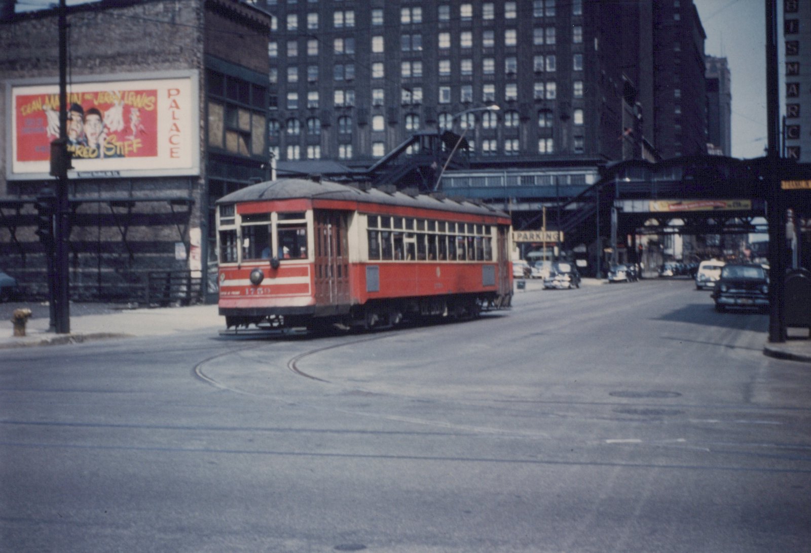 A red and cream streetcar, number 1759, turns on a city street with an elevated train structure overhead, surrounded by 1950s-era cars and a large movie billboard on a building.