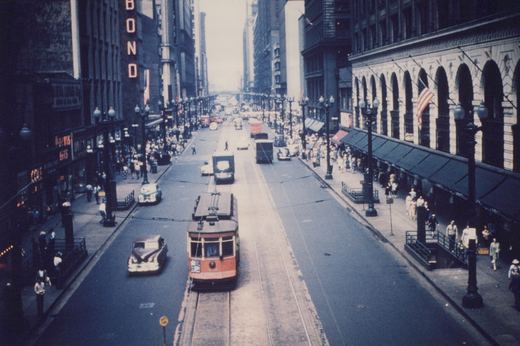 An overhead view of a red and cream Peter Witt streetcar on a wide city street with vintage automobiles and crowds of pedestrians on the sidewalks.