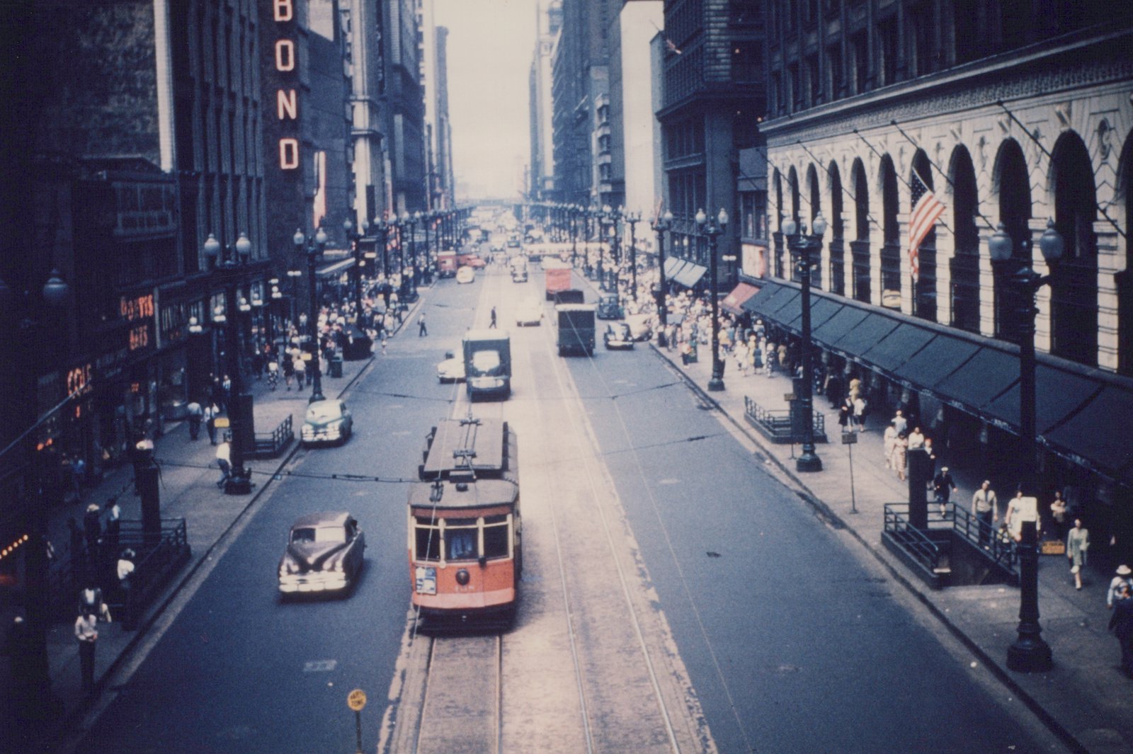 An overhead view of a red and cream Peter Witt streetcar on a wide city street with vintage automobiles and crowds of pedestrians on the sidewalks.