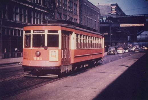A vintage orange and cream streetcar, numbered 531, runs down a city street under an elevated railway, with 1940s cars in the background.