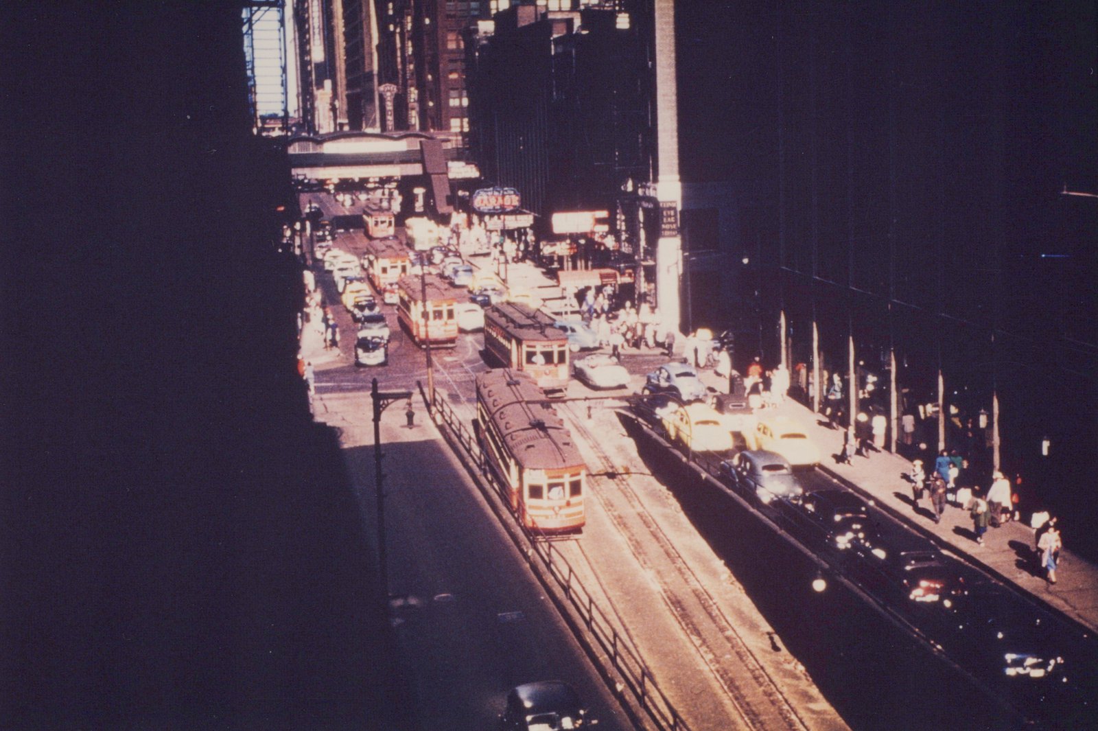 A high-angle, sunlit street scene in Chicago with multiple yellow and cream streetcars running on tracks between rows of cars and pedestrians on the sidewalks.