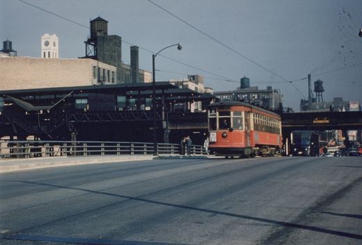 A vintage red streetcar labeled number 6435 and route 8 sits on tracks beside an elevated train station, with several people nearby and city buildings in the background.