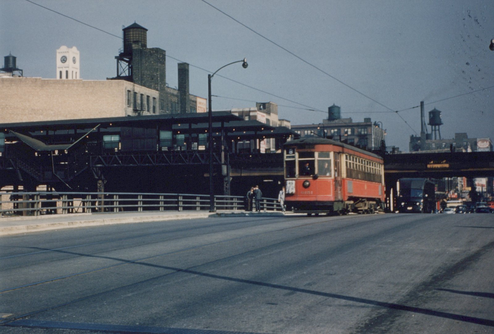 A vintage red streetcar labeled number 6435 and route 8 sits on tracks beside an elevated train station, with several people nearby and city buildings in the background.