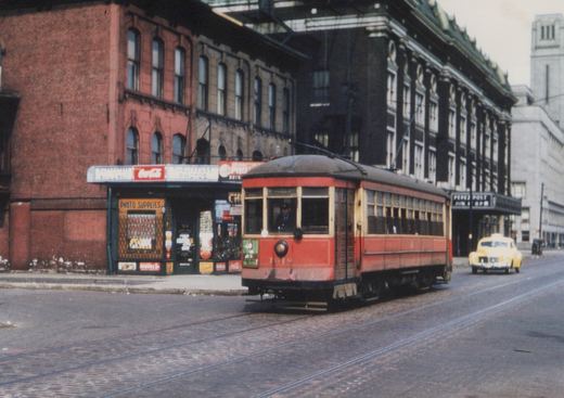 A vintage red streetcar with a curved roof runs on cobblestone tracks past a brick building with storefronts and a yellow car in the background.