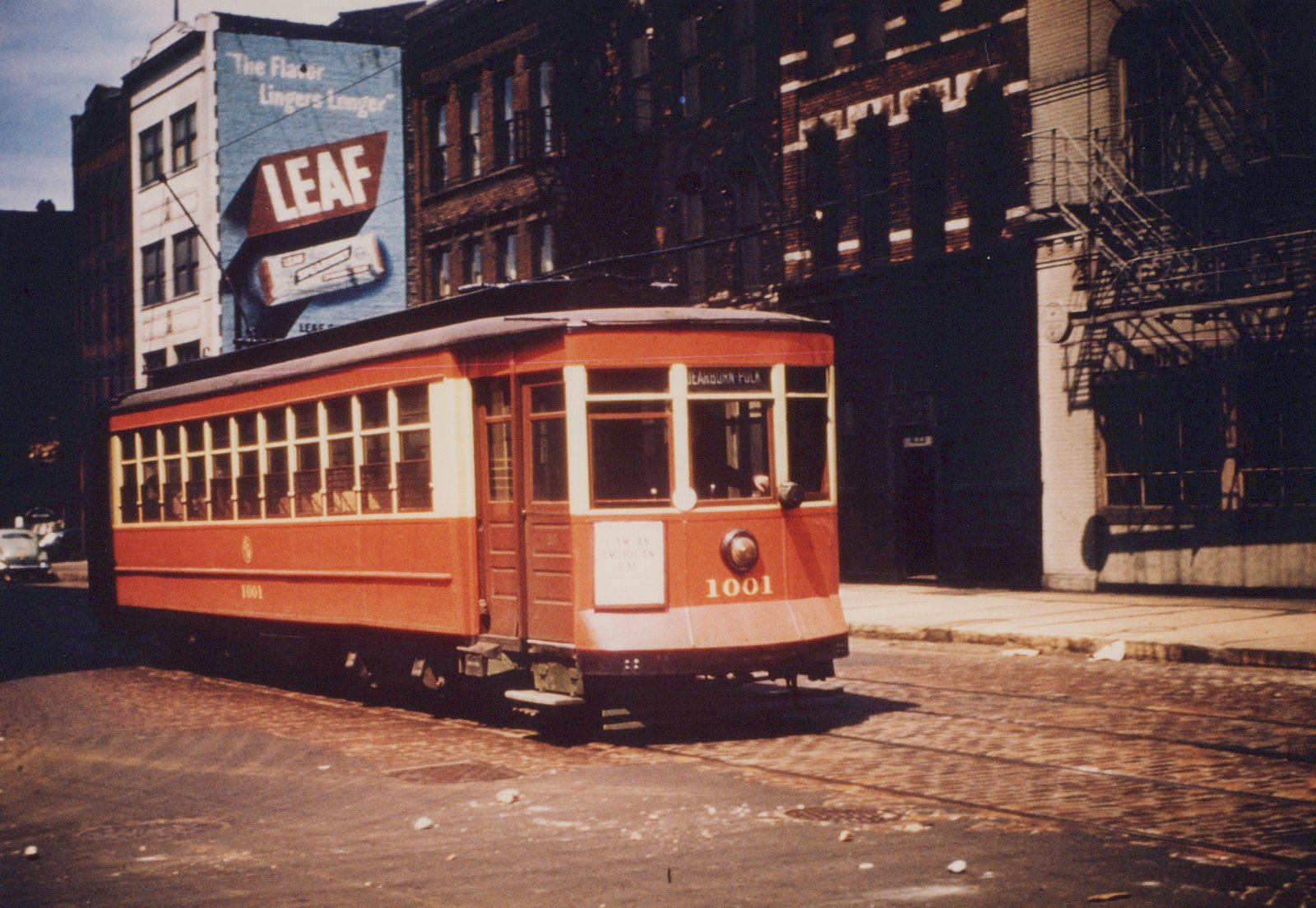 A red and cream Chicago Surface Lines streetcar numbered 1001 is shown from a three-quarter front view on a city street, with a large billboard for 'LEAF' cigarettes on a building behind it.