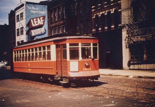 A red and cream Chicago Surface Lines streetcar numbered 1001 is shown from a three-quarter front view on a city street, with a large billboard for 'LEAF' cigarettes on a building behind it.