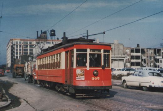 A vintage red streetcar with the number 565 on its front, shown at an angle on a city street with cars parked nearby and buildings in the background.