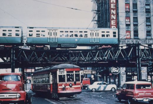 A red and cream streetcar with the number 1788 is on a city street, with a green and white elevated train passing overhead on a steel structure, surrounded by 1950s-era cars and storefronts.