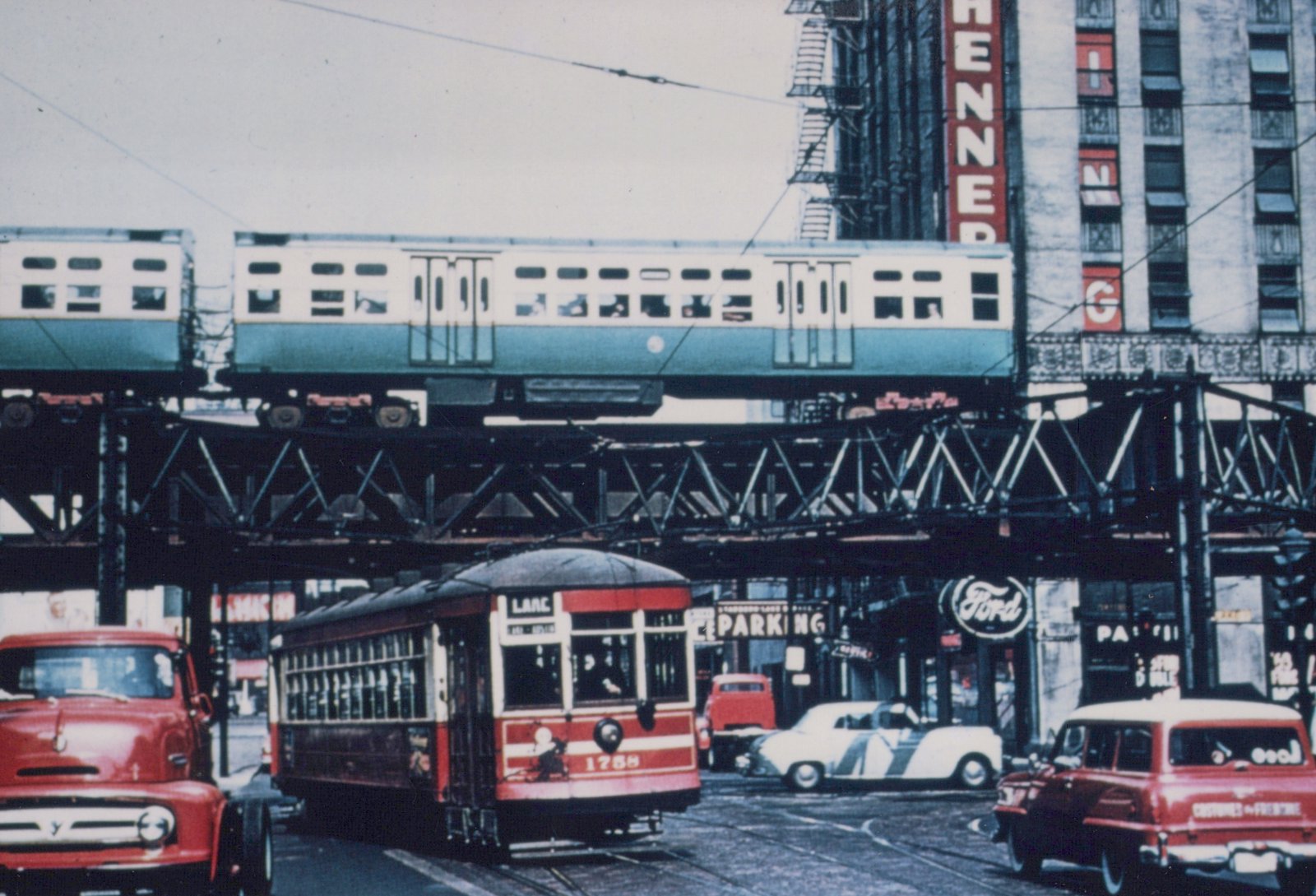 A red and cream streetcar with the number 1788 is on a city street, with a green and white elevated train passing overhead on a steel structure, surrounded by 1950s-era cars and storefronts.