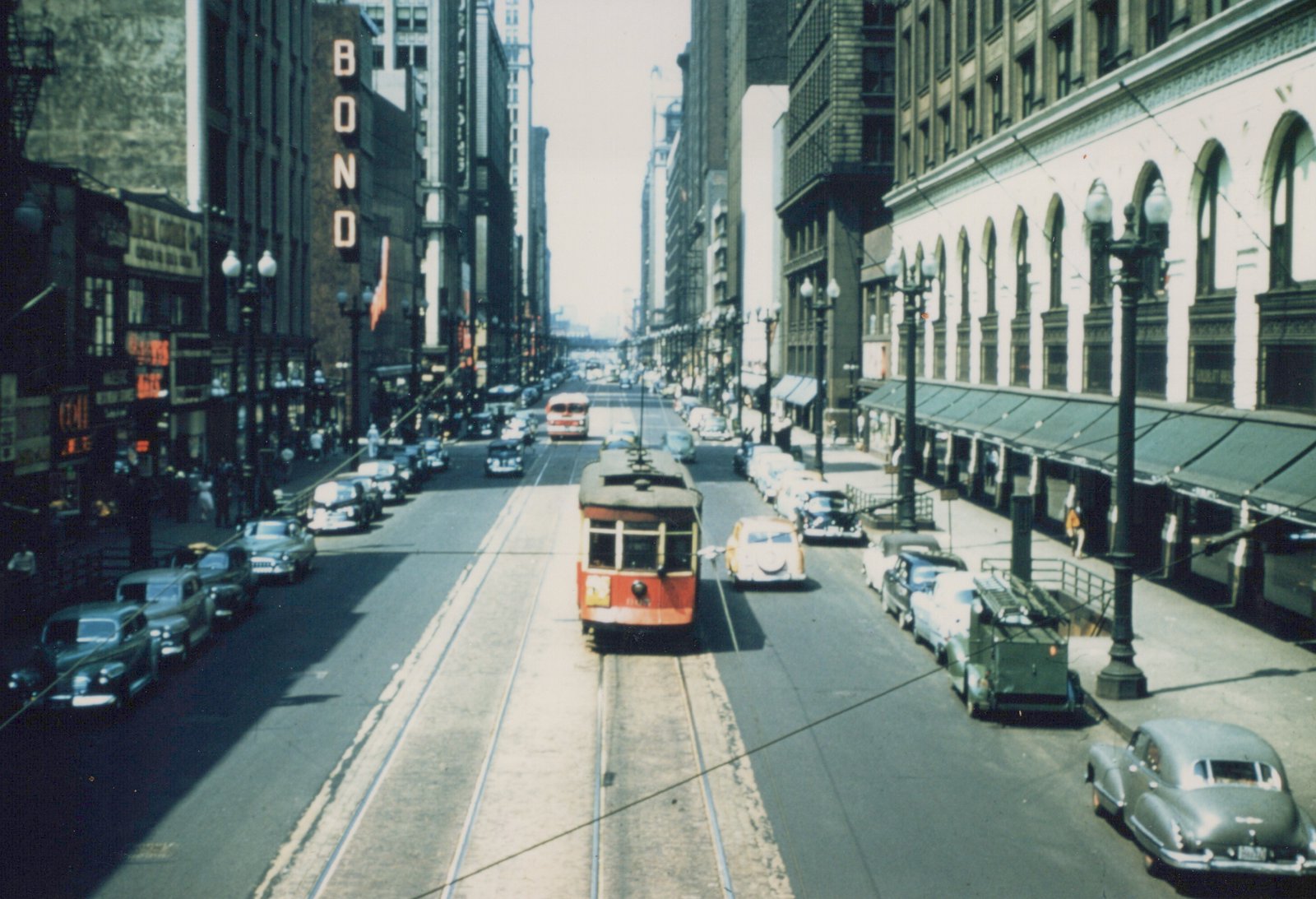 A red and cream streetcar travels down a busy urban street lined with vintage cars and tall buildings under a clear sky.