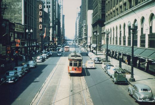 A red and cream streetcar travels down a busy urban street lined with vintage cars and tall buildings under a clear sky.