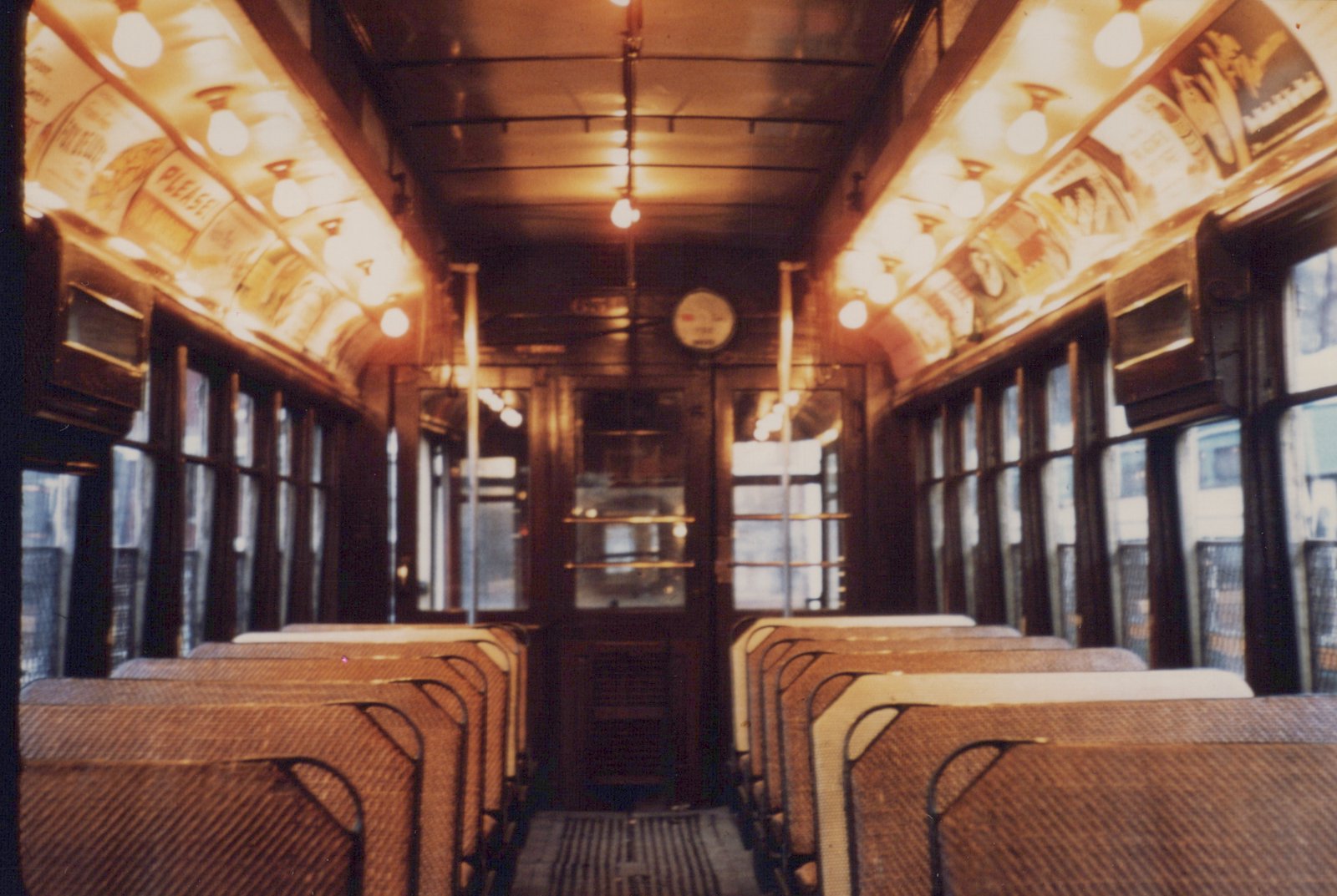 The interior of an old streetcar with wooden walls, woven seats, and overhead light bulbs illuminating vintage advertisements.