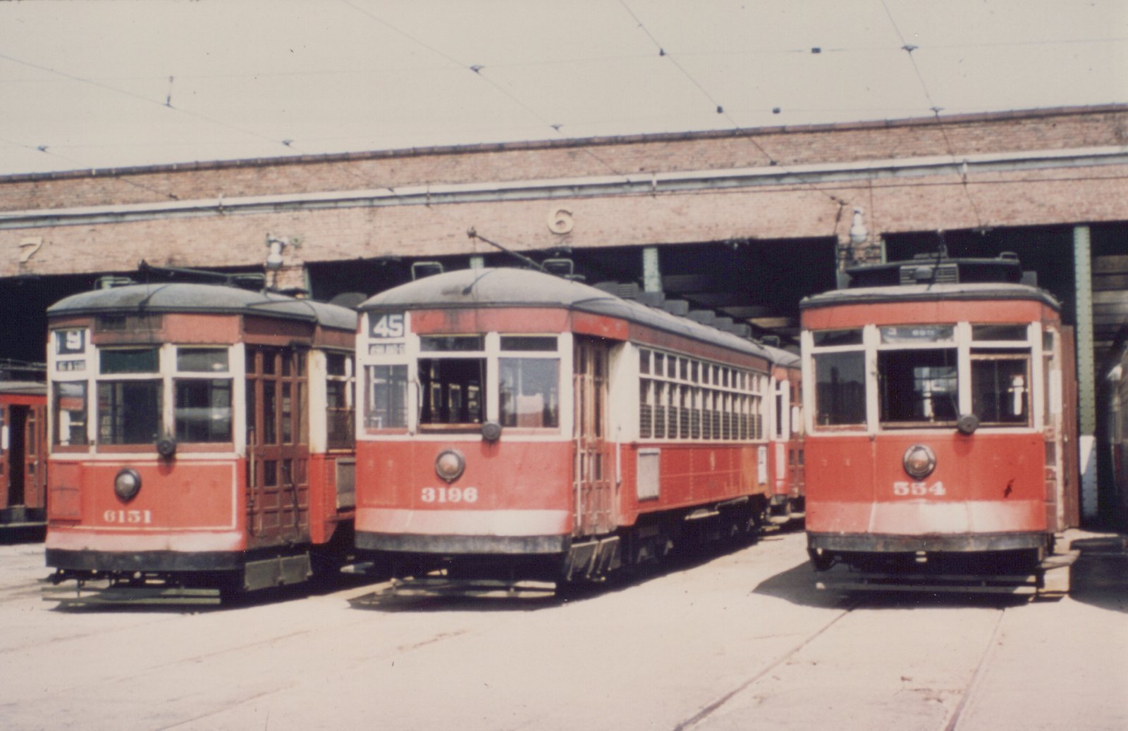 Three vintage red and white streetcars parked in front of a brick carhouse on a sunny day.