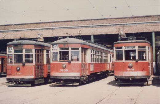 Three vintage red and white streetcars parked in front of a brick carhouse on a sunny day.