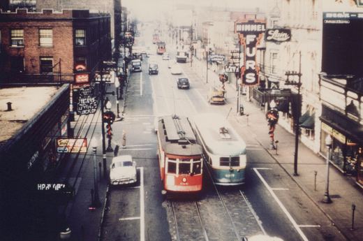 An elevated view of a city street in winter with two vintage streetcars in the foreground, one red and one green, surrounded by cars and brick buildings with signs.
