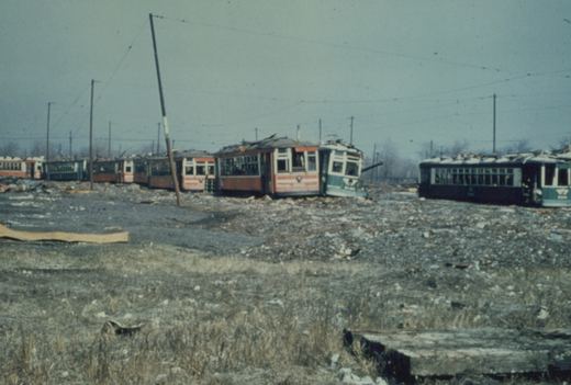 A row of old, damaged interurban railcars with green and cream livery sit in a gravel and debris-filled scrapyard under overhead wires.