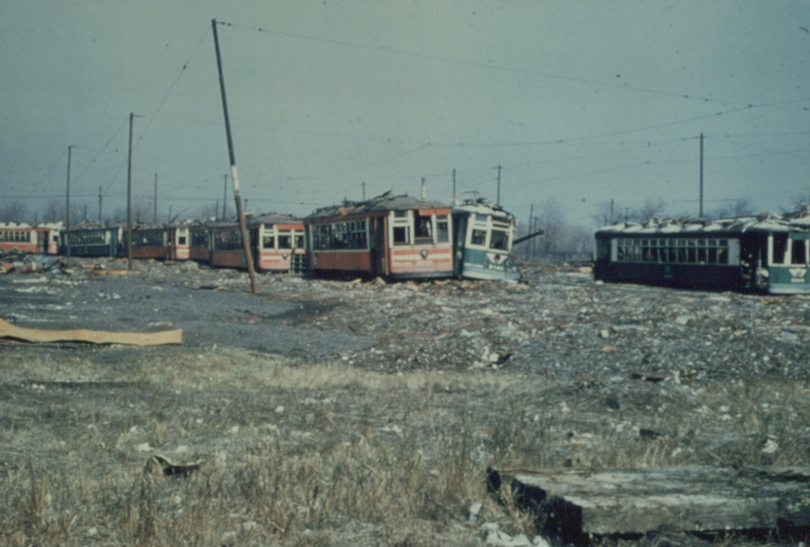 A row of old, damaged interurban railcars with green and cream livery sit in a gravel and debris-filled scrapyard under overhead wires.