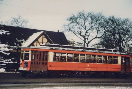 A red and cream interurban car with the number 505 on its side, labeled 'MILWAUKEE' on its front, is shown on a snowy street with passengers visible through its windows, in front of a Tudor-style building.