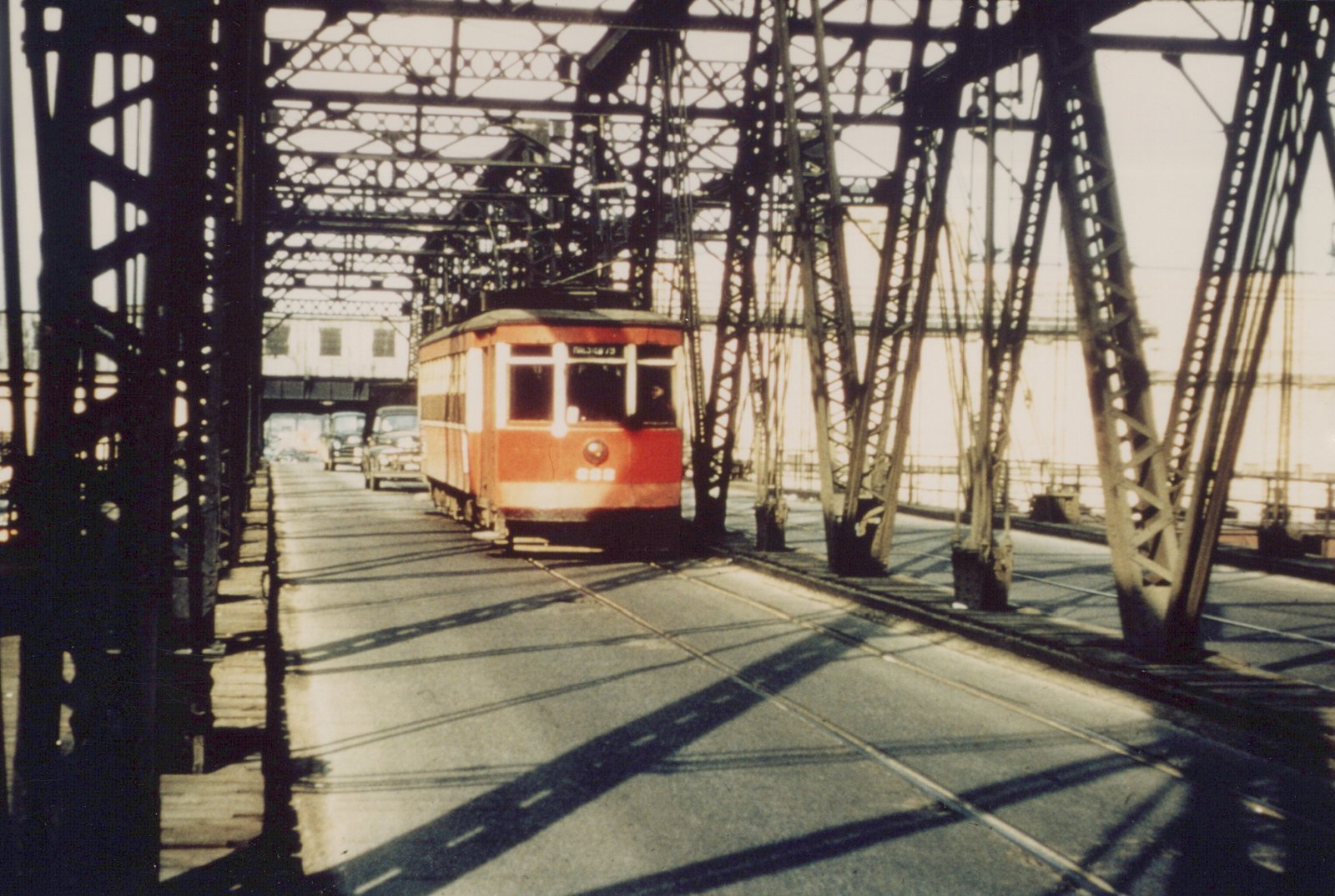 A vintage red streetcar numbered 688 travels across a metal truss bridge, with its destination sign showing 63RD-STONY, and cars following behind under bright sunlight.