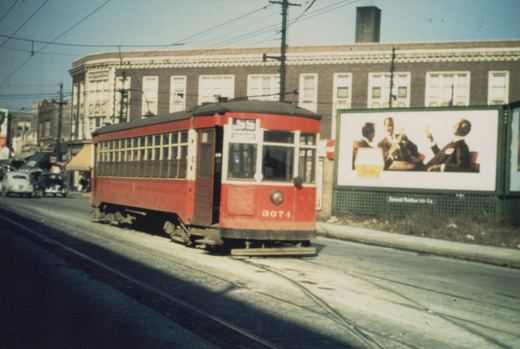 A red Chicago Surface Lines streetcar, number 3074, travels down a city street with overhead wires, past a large billboard and older automobiles.