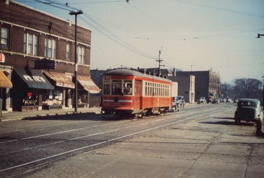 A red and cream streetcar numbered 4181 travels down a paved street past storefronts, including one named Pete's, with vintage automobiles parked along the sides.