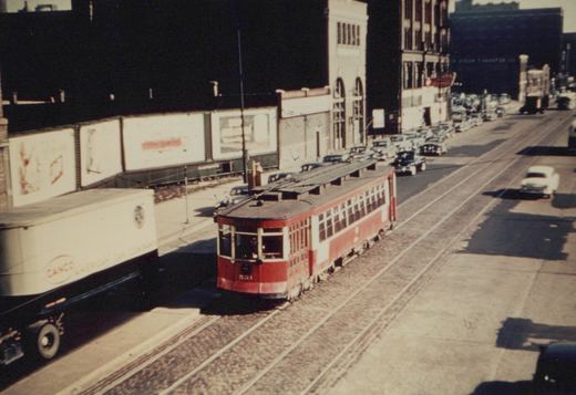 A vintage red Chicago streetcar with the number 551 travels down a busy urban street lined with parked cars and a white delivery truck.