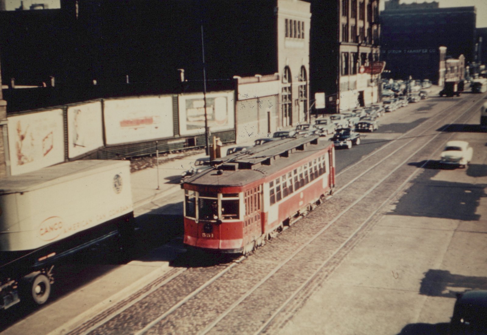 A vintage red Chicago streetcar with the number 551 travels down a busy urban street lined with parked cars and a white delivery truck.